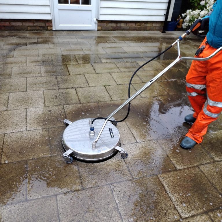 A person using a pressure washer on a patio, with flowers and garden furniture in the background.